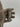 A silver metal watch band with a central clasp engraved with a logo, held by a hand wearing a beige glove against a light gray background.